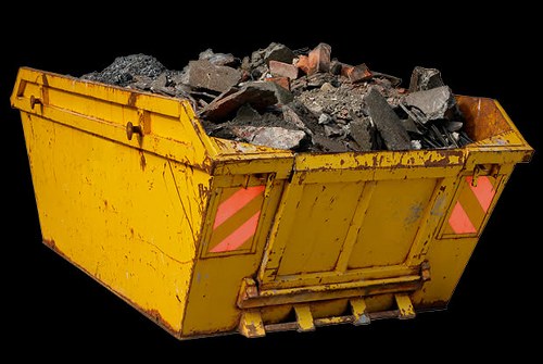 A large, yellow industrial skip filled with broken concrete, bricks, and gravel, positioned outdoors on a paved surface against a black background. The skip's metal sides display visible wear and rust spots, with pink reflective safety markings on the rear. The debris inside varies in size and texture, with rough, jagged edges and dark grey and reddish hues. The skip is partially elevated at the front, showing the lifting mechanism at the bottom. The scene appears to be part of a commercial waste removal operation likely serving a building or construction site in Ilford, with the company Commercial Waste Removal Ilford providing rubbish collection services.