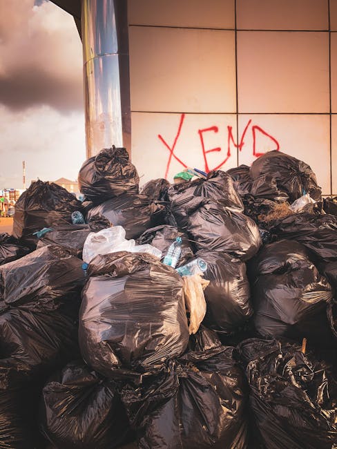 A large pile of black rubbish bags, some partially open revealing plastic bottles and discarded waste, is situated outdoors in front of a modern building with tiled exterior walls. The bags are stacke