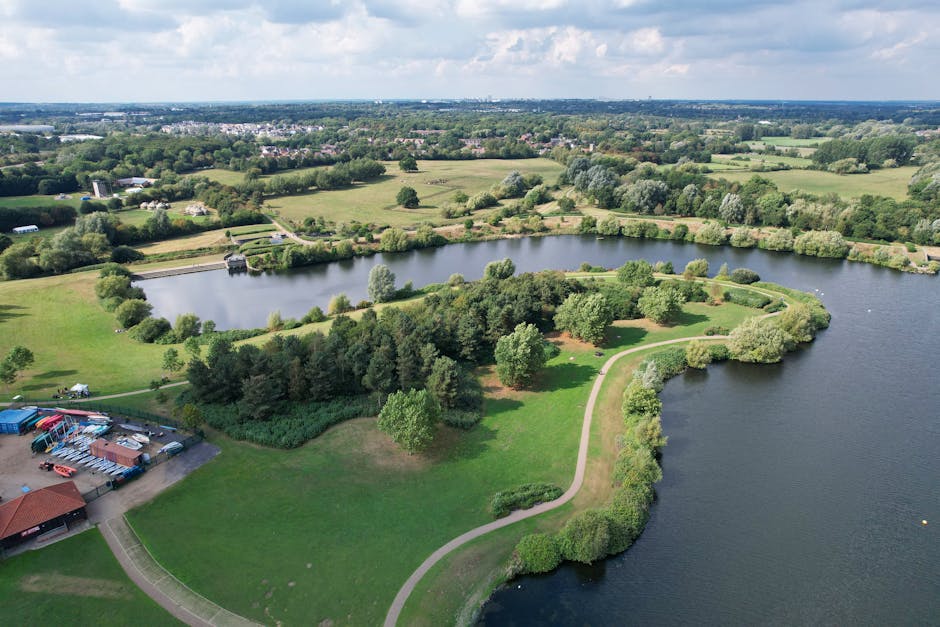 Aerial view of a large, scenic park featuring a winding pathway that runs along the edge of a calm, dark blue lake, shaped in a semi-circular peninsula with lush green trees and shrubs lining its peri
